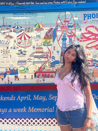 Young person with long dark hair wearing a pink lace-trimmed camisole and denim shorts, smiling slightly while standing in front of a colorful amusement park mural.
