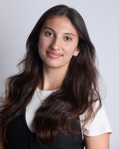A young person with long dark hair, wearing a white shirt and black vest, smiling slightly against a plain background.