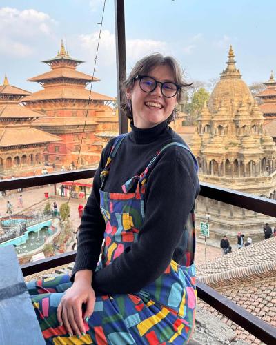 Smiling person wearing glasses, a black sweater, and colorful patterned overalls sits on a balcony. Behind them are historic temples with tiered pagoda-style roofs and stone carvings, set against a blue sky and a bustling courtyard below.