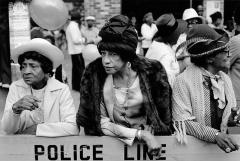 A monochrome photo of a person with a fur coat standing among a group of people, behind a barricade marked “Police Line”.