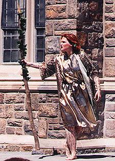 Photo of a performance of Bacchus in 1999 of a woman in costume holding a long stick in front of a stone building.