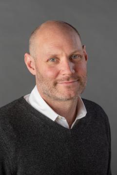 Hadley Creighton Bergstrom wearing a white collared shirt and dark gray sweater against a gray background.