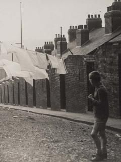 An old monochrome photo of a child in a street, with rows of brick buildings nearby.