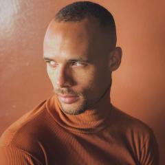 Head and shoulders photo of Mike Tyus wearing an orange shirt with a light orange background. 