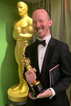 Jon Read wearing a tux and holding and Oscar statuette while squeezing his eyes shut and smiling; behind him is a large 3D replica of an Oscar award.