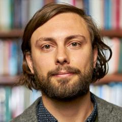 Headshot of Vincent Roy Hiscock wearing a gray jacket, navy patterned shirt and bookshelves in the background