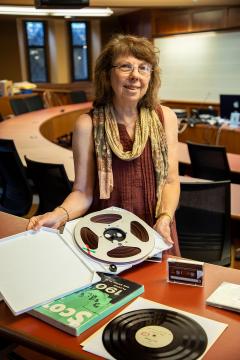 Woman wearing brown shirt and patterned scarf holding a reel-to-reel audio tape