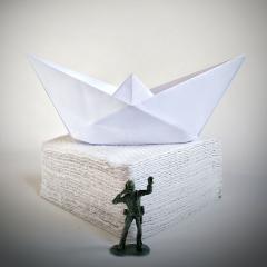 Paper boat sculpture - an army soldier figure standing in front of a white paper boat against a light background.