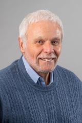 Sidney Plotkin wearing a light blue collared shirt and blue sweater against a gray background.