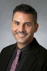 Carlos Alamo wearing a black shirt and red patterned tie against a gray background.