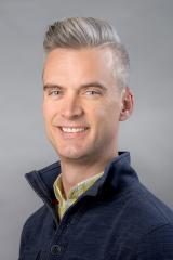 Colin Echeverría Aitken wearing navy shirt with high collar against a gray background.