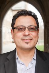 David Eduardo Tavarez wearing a light patterned shirt and black jacket in front of an archway.