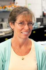 Kathleen M. Susman wearing a yellow shirt, light green jacket with lab equipment in the background.