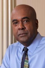 Luke C. Harris wearing a light blue collared shirt and multi colored tie against a light background.