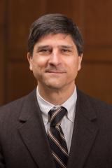 A person wearing a light colored shirt and brown jacket and tie smiles at the camera. They are in a room with wood paneling in the background.