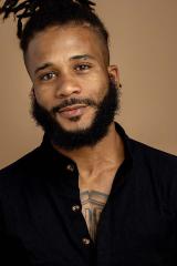 A photo of a person with dark, curly hair and beard, and a black shirt, looking at the camera