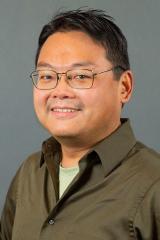 Benjamin Ho wearing a dark tan shirt against a dark gray background.