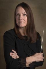 A person with long brown hair and a black formal shirt looks at the camera. The person is holding a white conductor's wand.