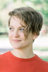 A portrait of a person with short blond hair and a red shirt, smiling and looking to the viewer's left. In the background is a blurry suggestion of foliage.