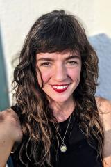 Woman with dark, curley hair, black dress, silver necklace, smiling for the camera.