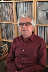 A man with dark glasses and gray hair sitting in front of a bookcase filled with vinyl records.