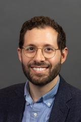 Headshot of person with dark curly short hair, glasses, and beard wearing a dark suit jack and light blue collared shirt.