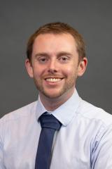 Person with short brown hair in a white collared shirt and blue tie smiling for a portrait,