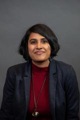 A person with medium length dark hair, dark skin, smiling, wearing a dark blue blazer, red mock-neck shirt, and long necklace with a gold pendant.