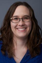 Portrait of a smiling person with long brown hair, glasses, and a blue shirt.