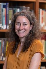 Person with long curly brown hair standing in front of a book case.