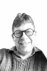 A grayscale portrait of a person wearing round glasses and a textured knit sweater, photographed in black and white against a bright background.