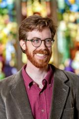 Portrait of a smiling individual in glasses, standing in front of a stained glass window as the backdrop.