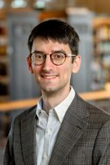Tommasi Pier Carlo smiling with glasses, wearing a brown plaid blazer and white shirt, with a blurred library background.