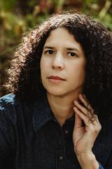 A person with dark, shoulder-length curly hair wears a dark blue denim button-up shirt and rests one hand against their neck while looking forward. The background is a softly blurred outdoor setting with natural sunlight.