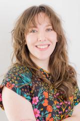 Maya Van Nuys with long wavy brown hair, bangs, and blue eyes, smiling warmly with arms crossed while wearing a black short-sleeved top with a vibrant colorful floral pattern.