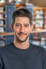 Jesse Delia smiling with short dark hair and light stubble, wearing a plain dark crewneck shirt in a library with wooden bookshelves blurred in the background.