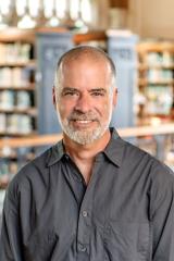 Chris Grove with short hair and a graying beard, wearing a dark gray button-up shirt set against a softly blurred library background with wooden bookshelves.