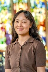 Soo Jin Jeon with long dark wavy hair, wearing a brown short-sleeved button-up knit shirt and a delicate necklace, set against a softly blurred background of a colorful stained-glass window.