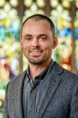 John Garrard Stehlin smiling with short dark hair, light facial hair, and blue eyes, wearing a textured gray blazer over a dark button-down shirt against a softly blurred stained-glass window background.