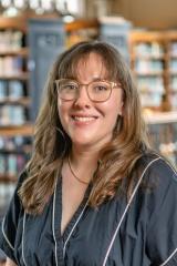 Maya Marie Van Nuys smiling, long wavy brown hair with bangs, and clear-framed glasses, wearing a black top with white piping in front of a softly blurred library background with wooden bookshelves.