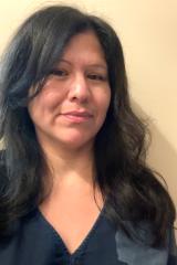 Leora Gansworth with long dark wavy hair, wearing a dark V-neck top and smiling gently against a light neutral wall.