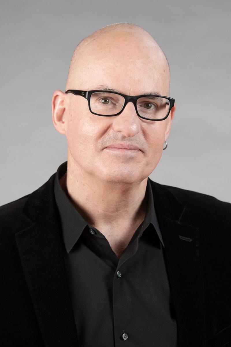 Michael J. Walsh with glasses and a black shirt/blazer, against a gray background.