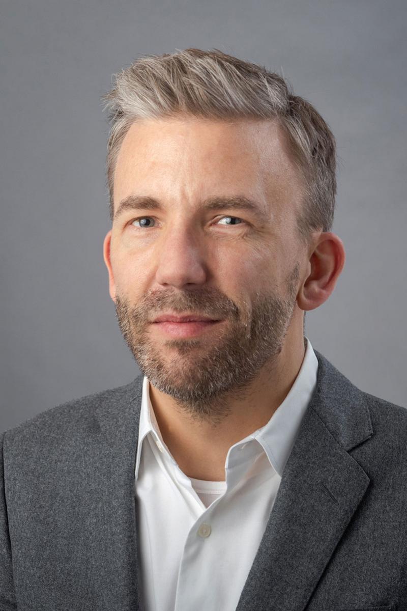 Tobias Armborst wearing a white collared shirt and dark gray jacket against a gray background.