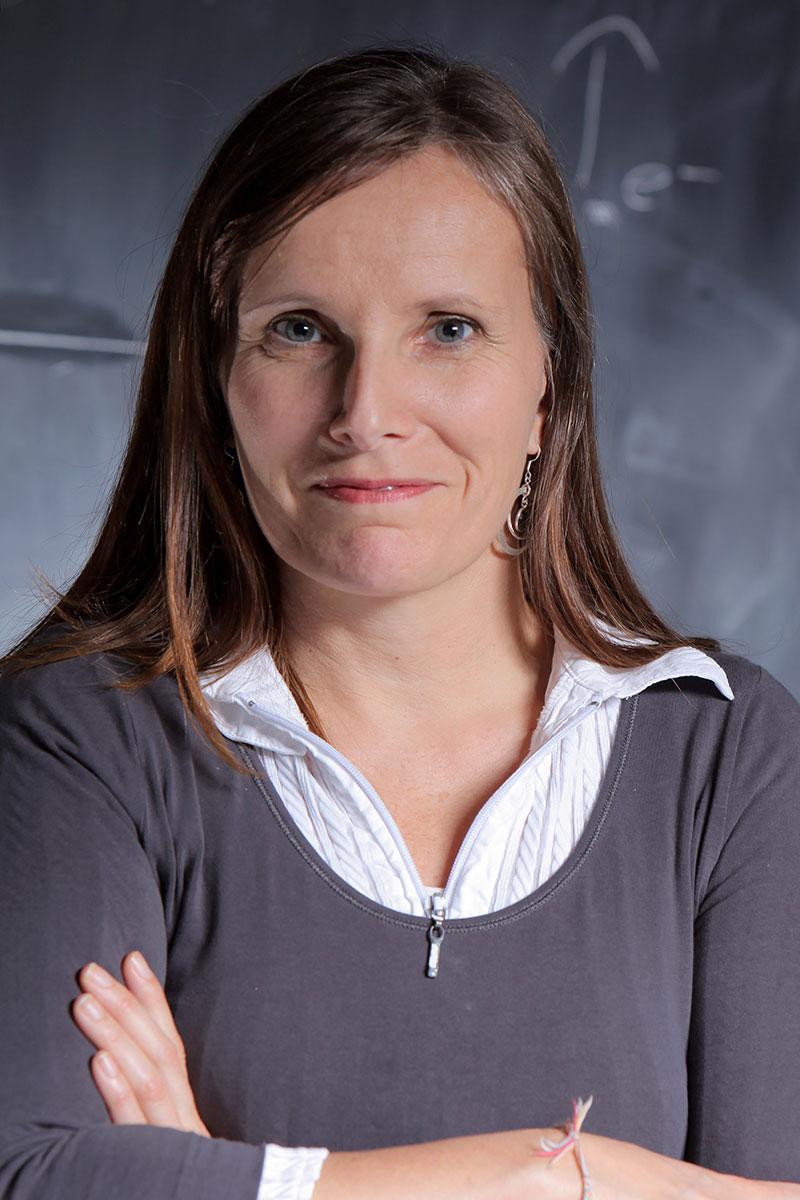 Jenny Magnes with long brown hair standing with arms crossed, wearing a white collared shirt under a gray sweater, posing in front of a chalkboard.
