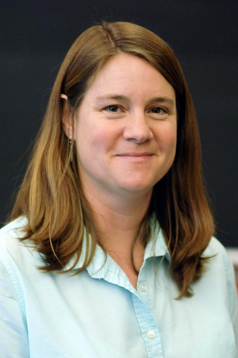 Jodi Schwarz smiling with long brown hair, wearing a light blue collared shirt against a dark background.