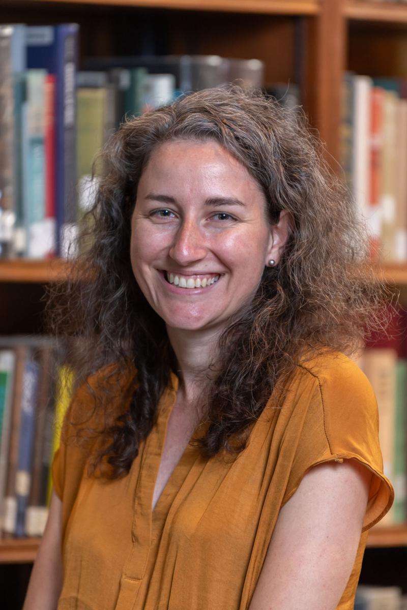 Person with long curly brown hair standing in front of a book case.