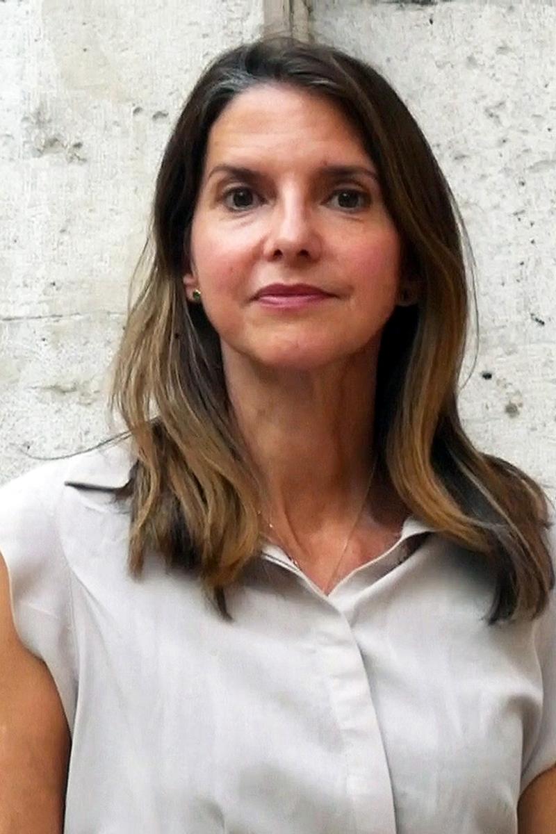Eva Woods Peiró with straight brown hair wearing a light gray blouse, standing against a textured stone wall.