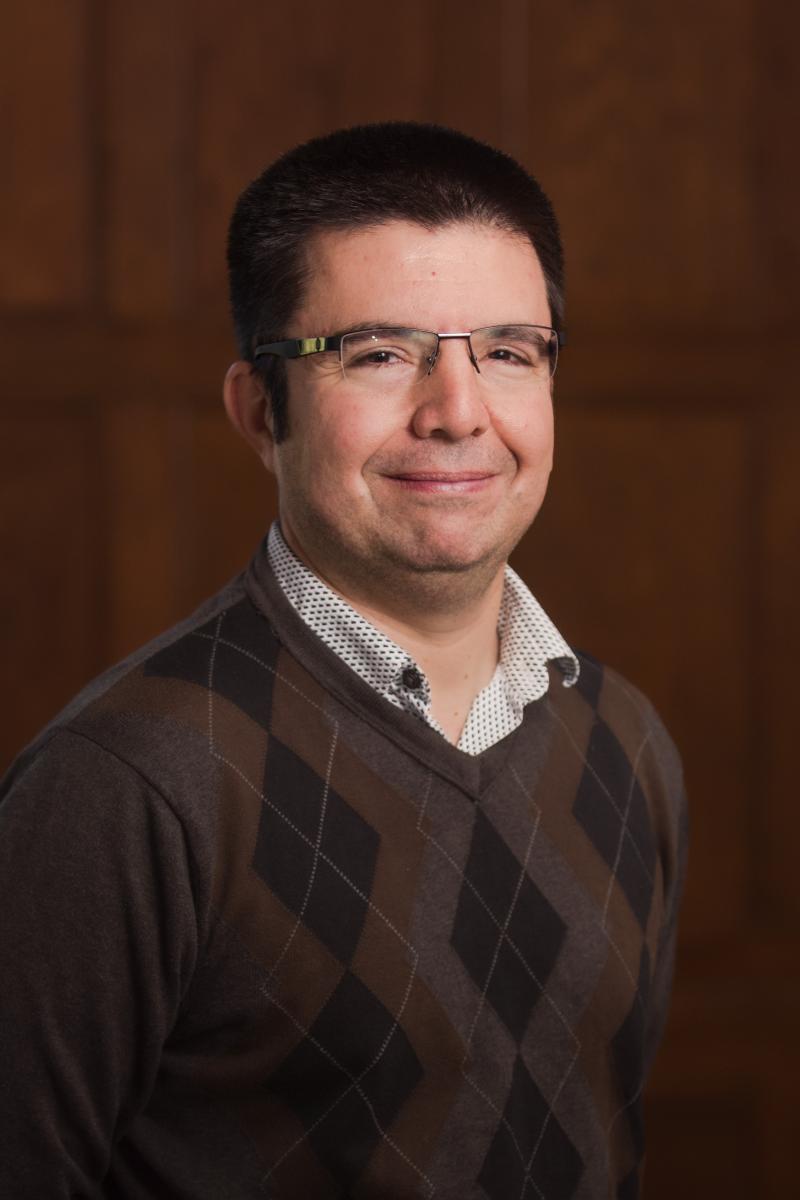Portrait of a person with short dark hair and glasses, dressed in a brown argyle sweater over a white button-down shirt, standing against a paneled wood background.