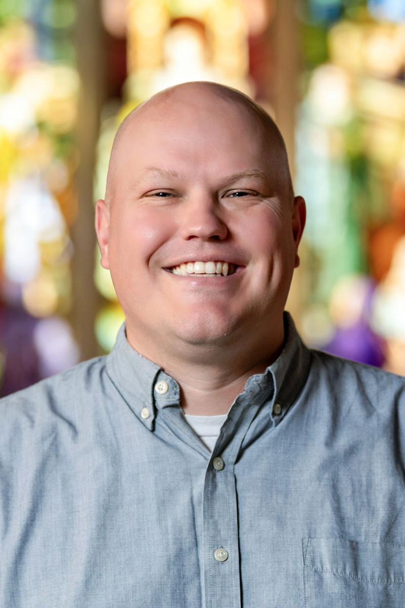 Portrait of a person with a bright smile, standing in front of a colorful stained glass window.