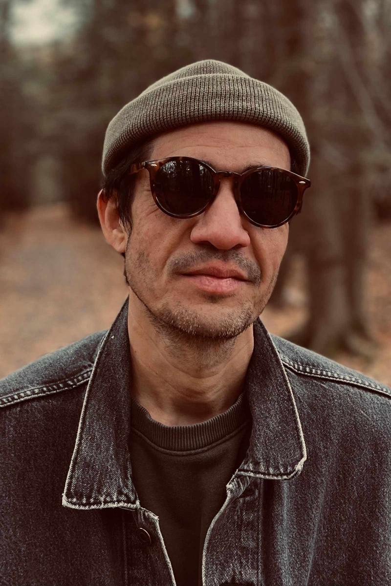 A person outdoors, wearing a dark denim jacket, a dark olive beanie, and round, tortoiseshell sunglasses. The background is a blurry, wooded area with brown and tan tones.